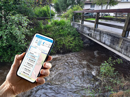 Es wird ein Handy gehalten, auf dem das NIVUS WebPortal abgebildet ist. Diese zeigt die Messdaten des Flusses im Hintergrund. Hier ist ein Sensor an einer befahrbaren Brücke installiert.